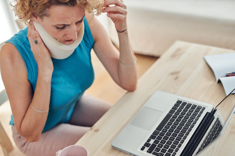 Woman with a Neck Brace Sits at a Wooden Desk, Holding Her Neck in Discomfort While Looking at an Open Laptop Nearby.
