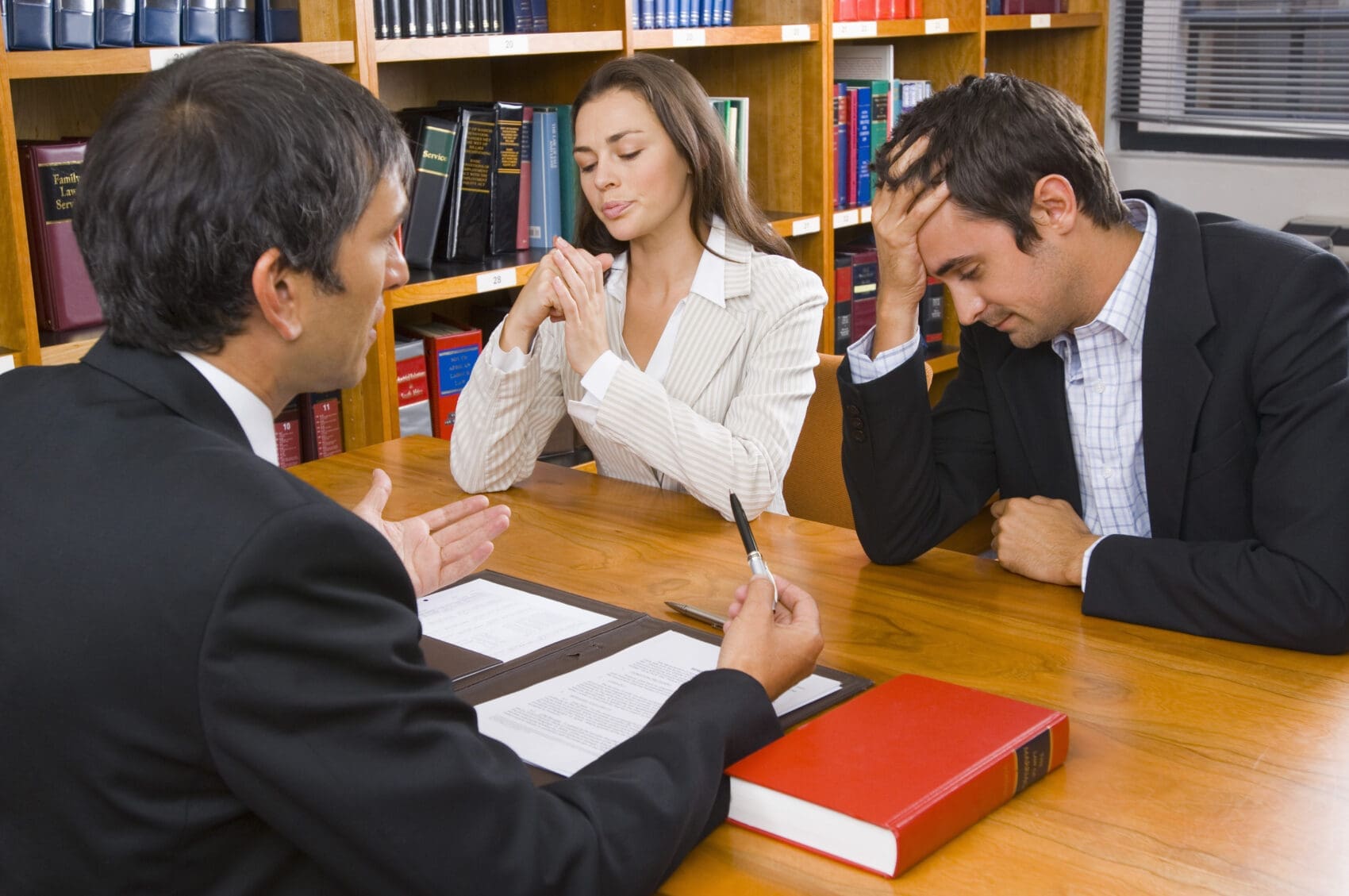 Lawyer Explains Documents to a Distressed Couple in a Library, Seated at a Wooden Table with Legal Books Nearby.
