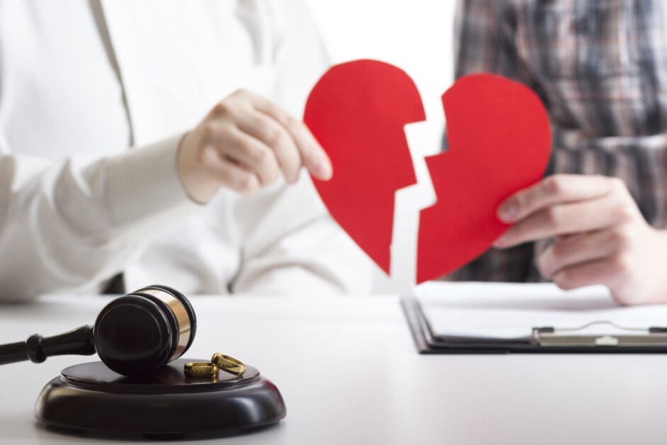 Two People Hold a Broken Red Heart Between Them, with a Judge's Gavel and Wedding Rings on a Desk, Suggesting Divorce or Legal Separation.