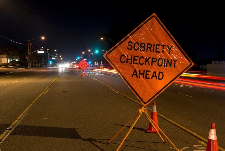 Nighttime Street Scene with an Orange Sobriety Checkpoint Sign and Traffic Cones Blocking a Lane Ahead of a Road Closure