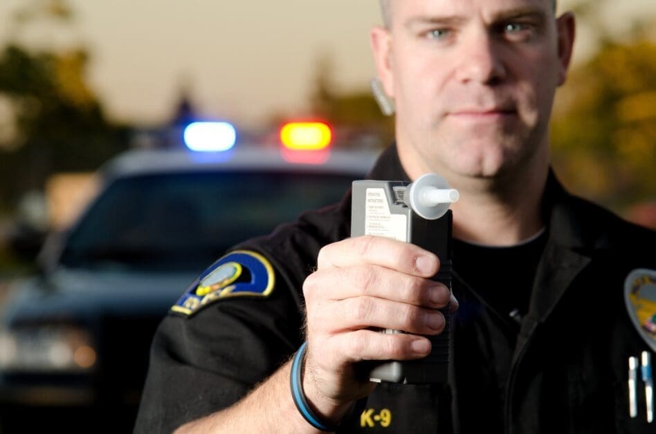 Close-up of a Police Officer in Uniform Holding a Handheld Device with a Nozzle, Police Car with Flashing Lights Behind Him.