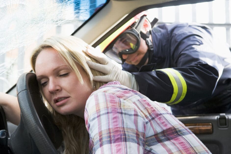 Distraught Woman Driver Grips the Steering Wheel As a Firefighter in a Helmet and Gloves Attends to Her Inside a Car with a Cracked Windshield, Emergency Scene.