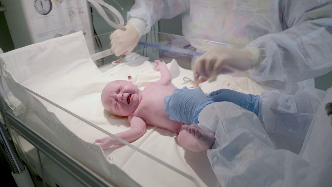 - Attorneys.Media - Legal Expert Interviews & Trusted Advice Crying Newborn Baby in a Neonatal Incubator with Medical Staff in Protective Gowns Nearby, a Blue Measuring Tape Across the Bed.