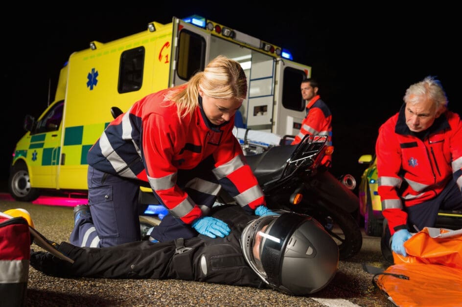 Paramedics in Red Jackets Attend to a Motorcyclist on the Ground at Night As an Ambulance Waits Behind Them.
