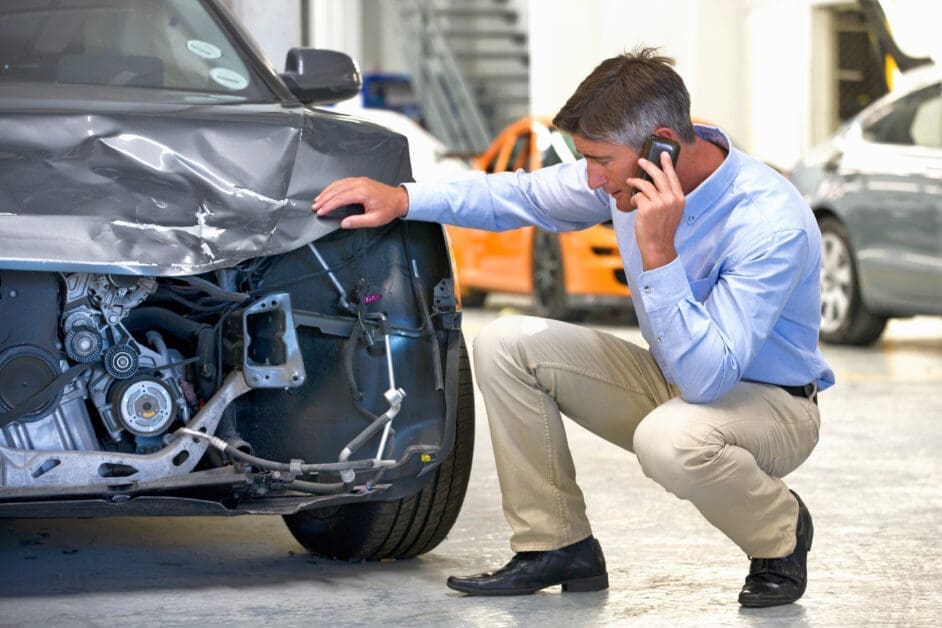 Male Mechanic Kneels Beside a Damaged Car Front, Talking on a Cellphone Inside a Repair Shop