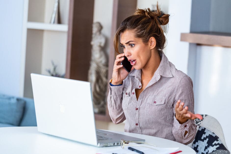 Woman Sitting at a Desk, Talking on a Smartphone While Looking at an Open Laptop with a Surprised Expression.