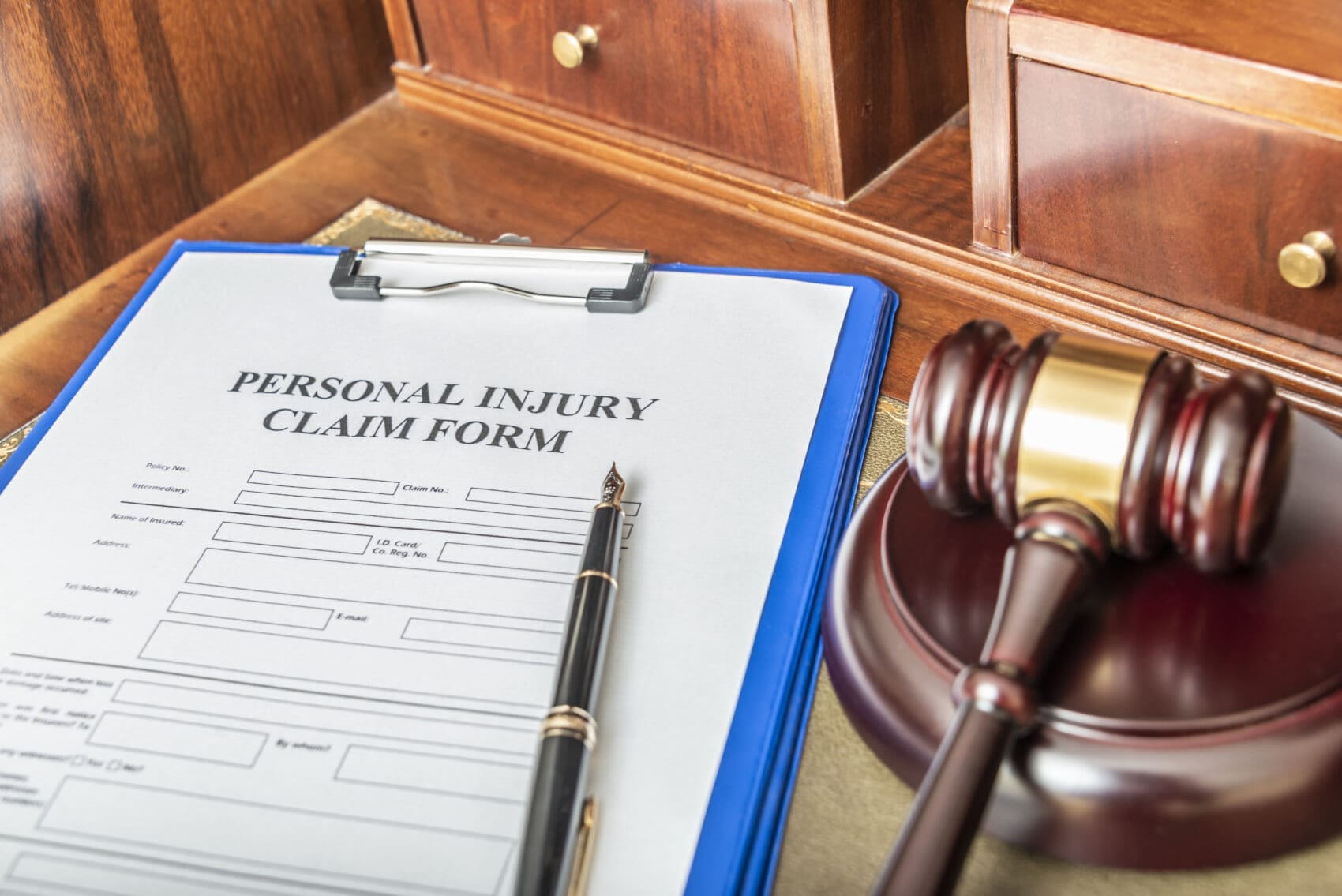 Personal Injury Claim Form on a Blue Clipboard with a Pen, Beside a Wooden Judge's Gavel on a Polished Desk.