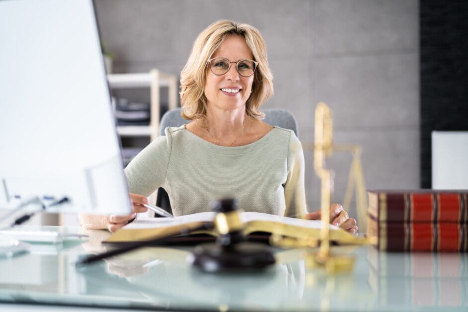 Smiling Professional Woman with Glasses Sits at a Glass Desk Reviewing Documents, with Law Books and a Gavel in the Foreground.