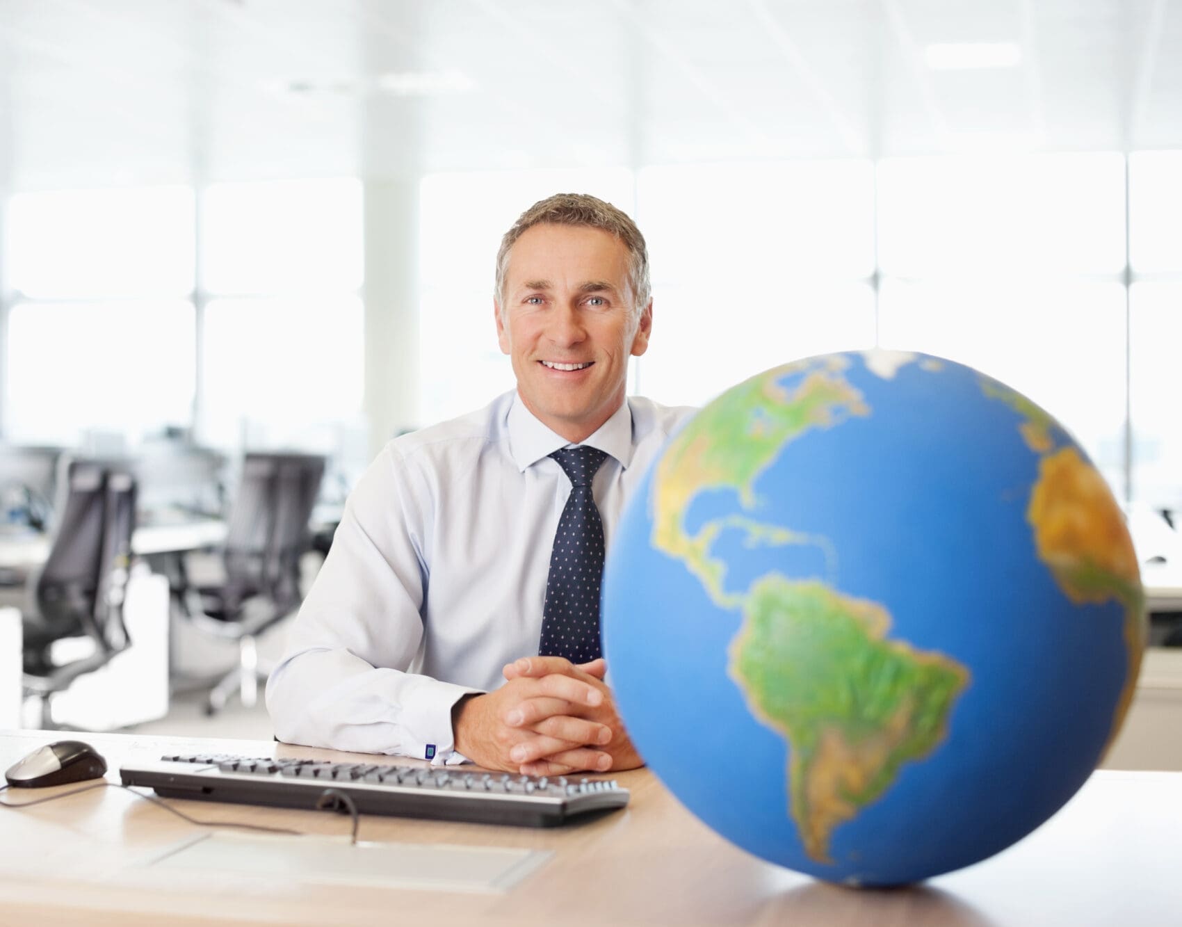 Smiling Man in a Shirt and Tie Sits at a Desk with a Large Globe in the Foreground, Office Background Behind Him.