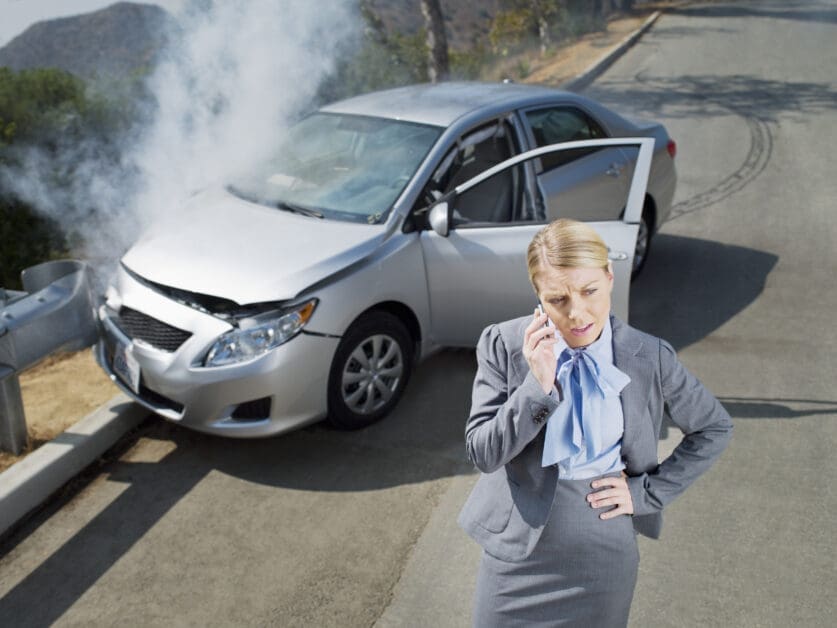 Smoldering Silver Sedan with Front-end Damage on a Winding Road; a Woman in a Gray Suit Stands Nearby and Talks on Her Phone.