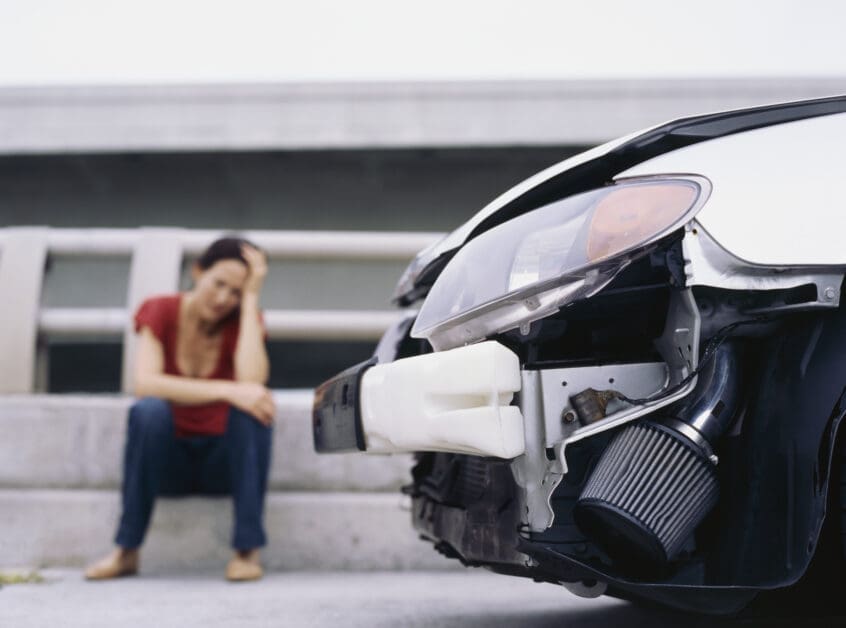 Woman Sitting on Steps, Head in Hand, Beside a Badly Damaged Car with Its Front End Exposed.