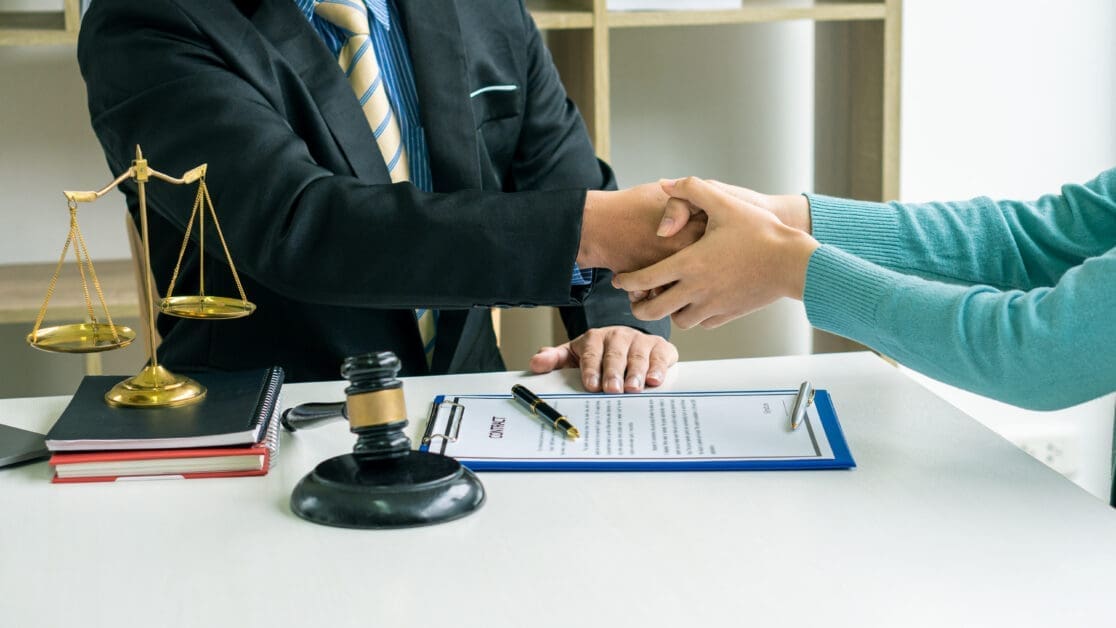 Handshake Across a Desk with Legal Documents, Scales of Justice, and a Gavel on the Table.