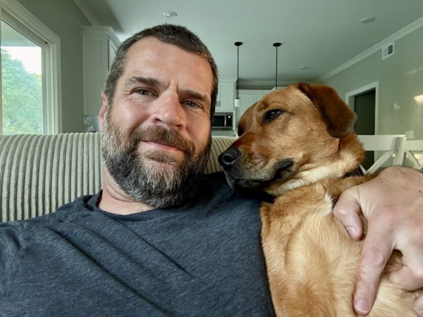 Man with a Beard Smiling While Snuggling a Brown Dog on a Striped Sofa in a Bright Home Interior.
