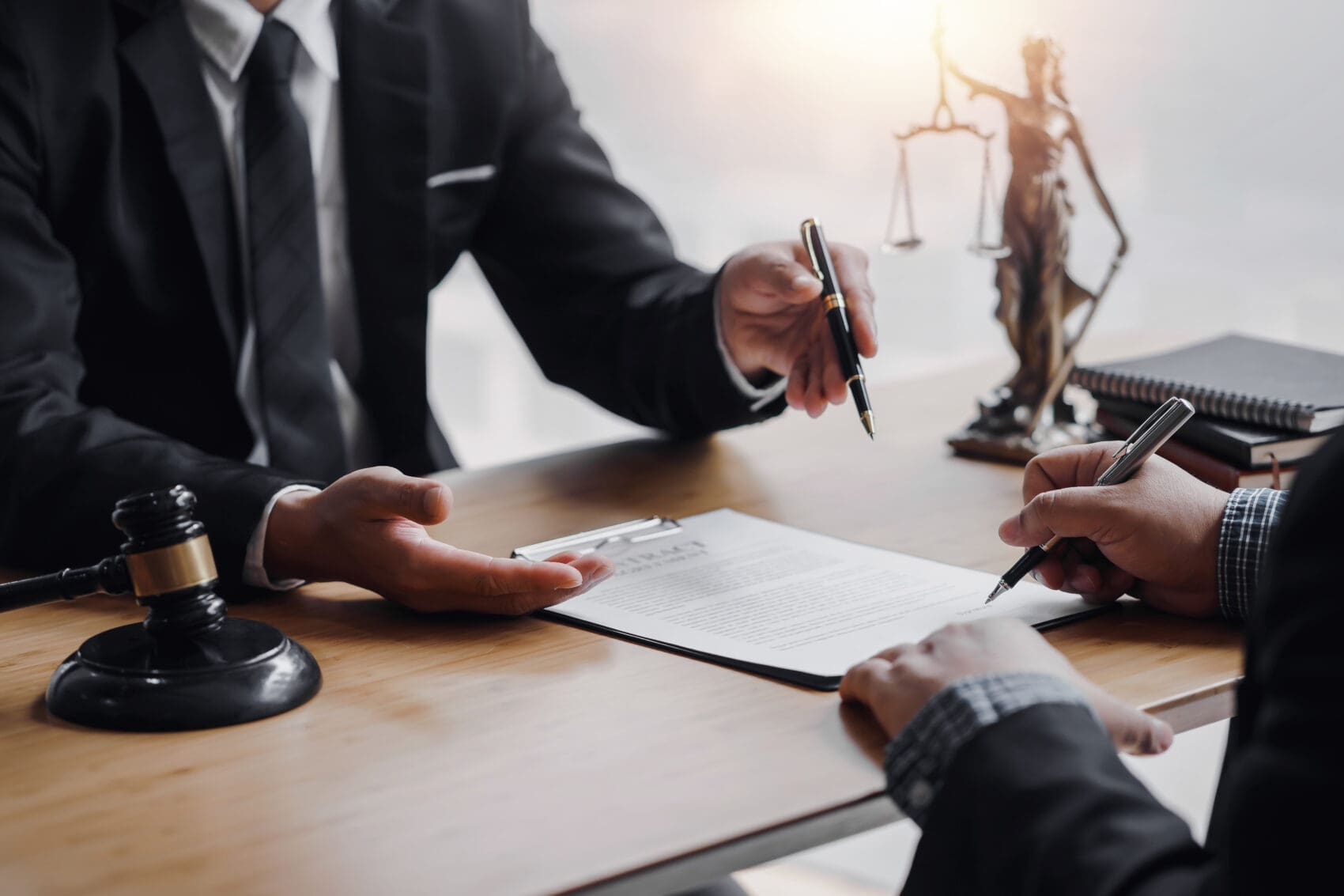 Two Professionals Sign a Contract at a Desk, One Holding a Pen and the Other Guiding with a Document, Legal Symbols in the Background