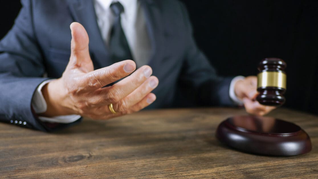 Gavel on a Wooden Table with a Suited Person Gesturing with an Open Hand Beside It, Signaling a Pause or Objection.