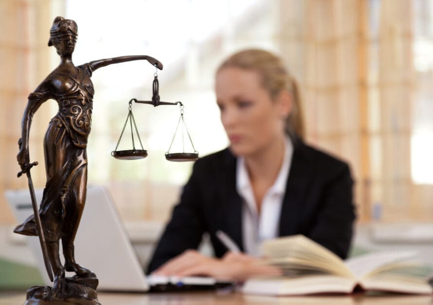 Statue of Lady Justice with Scales in Focus on a Desk; a Blurred Woman in a Suit Works at a Laptop in an Office Setting.