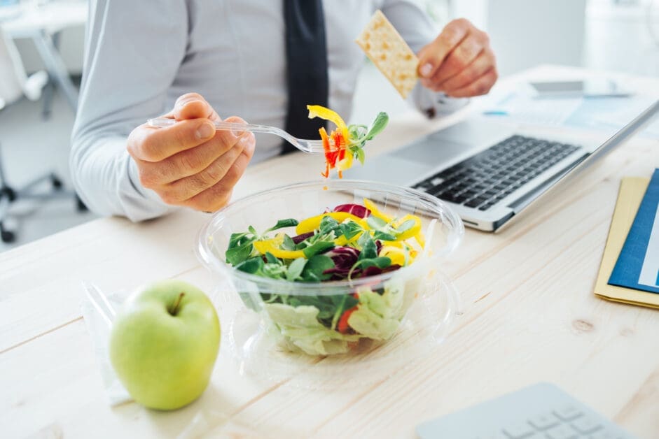 Person in a Shirt and Tie Eating a Colorful Salad at a Desk, Holding a Fork with Greens and Peppers While a Cracker is Held in the Other Hand, Laptop Nearby.