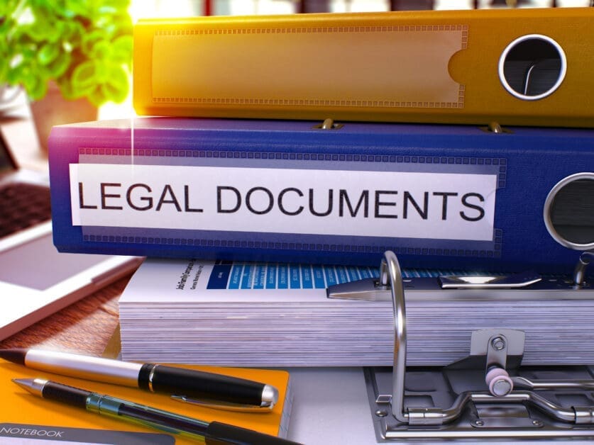 Blue Binder Labeled Legal Documents Stacked with a Yellow Binder on Top, on a Cluttered Desk with Papers and Pens
