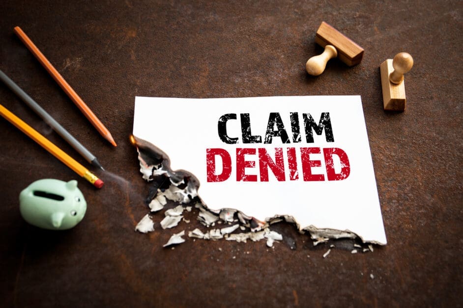 - Attorneys.Media - Legal Expert Interviews & Trusted Advice a White Paper Reading 'claim Denied' in Bold Black and Red, with Burnt Edges, on a Dark Brown Desk Beside Pencils and a Small Green Piggy Bank.