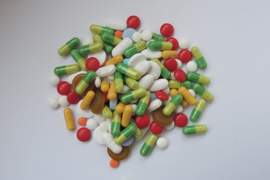 Pile of assorted pills and capsules in various colors scattered on a light surface, suggesting medication or supplement tablets mixed together.
