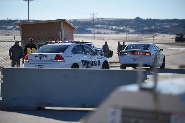 White Police Car with Blue Markings and Flashing Lights at a Roadblock; a Guard Booth and Several Officers in a Rural Desert Setting.