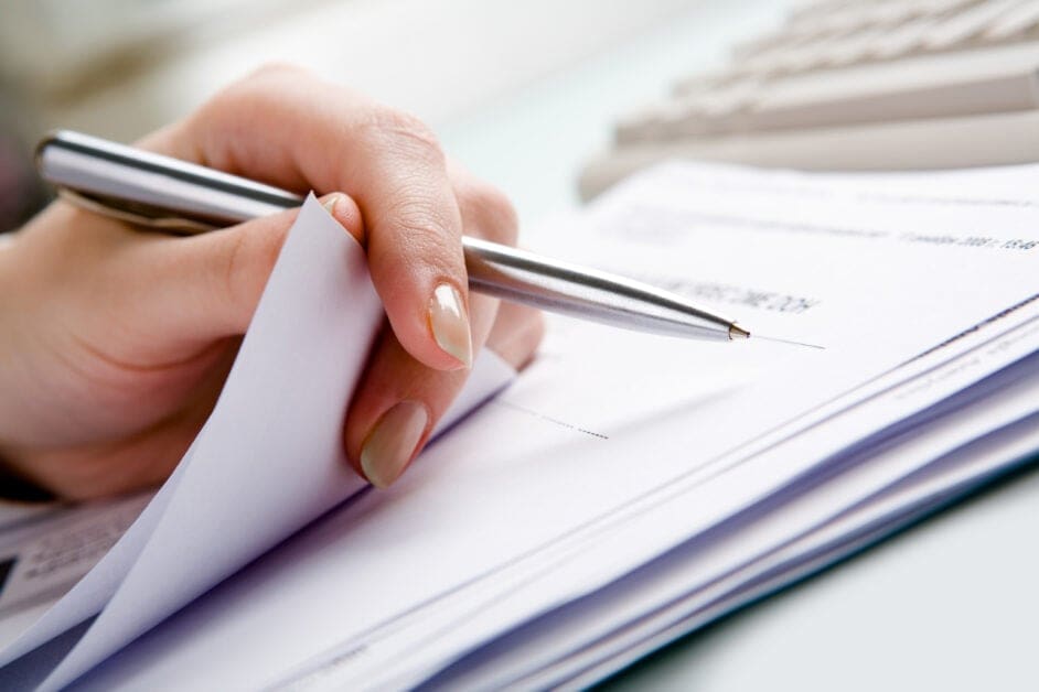 Close-up of a Hand Holding a Silver Pen and Signing a Stack of Papers on a Desk.