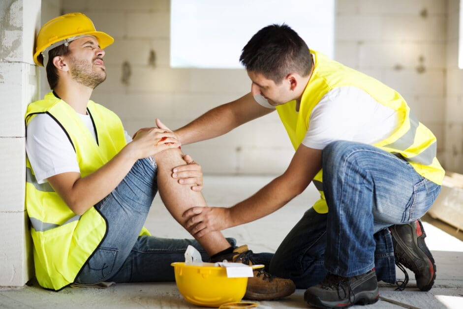 Two Construction Workers in High-visibility Vests; One Sits Against a Wall Clutching a Knee Injury While the Other Kneels to Help on a Building Site.