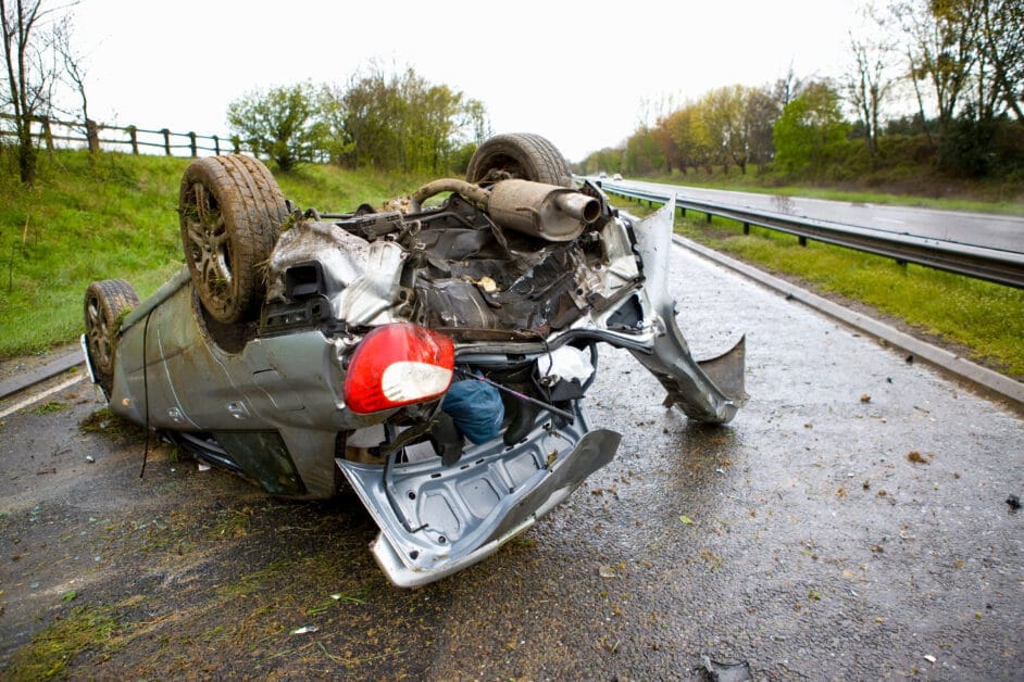 - Attorneys.Media - Legal Expert Interviews & Trusted Advice Car Flipped Onto Its Roof on a Wet Roadside, with a Damaged Rear and Exposed Undercarriage, Guardrail and Grassy Verge in the Background