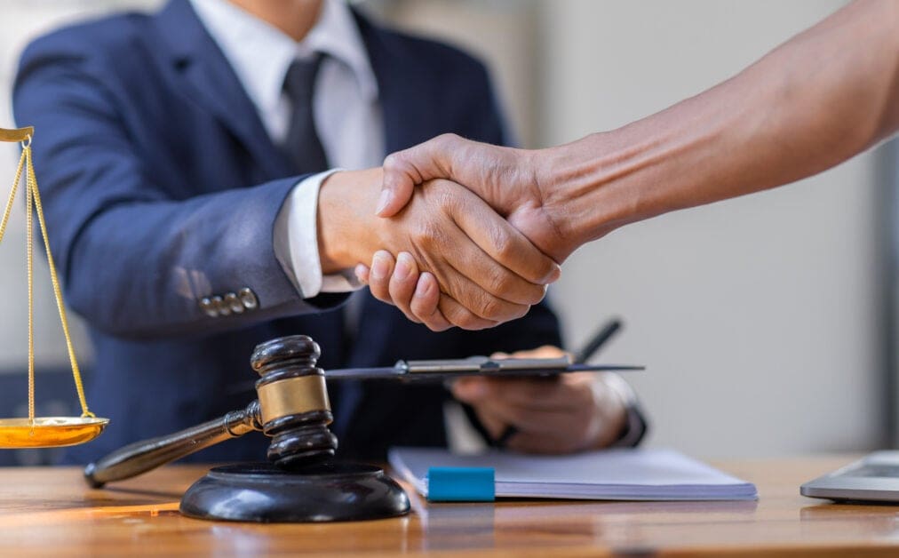 Handshake Between Two Professionals over a Desk with a Gavel and Scales, Symbolizing a Legal Agreement or Deal.