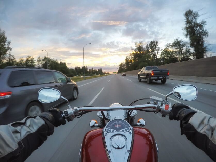 View from a Motorcycle Rider on a Multi-lane Highway at Dusk; Hands on Handlebars, Other Vehicles Ahead and to Sides, Sky with Clouds.