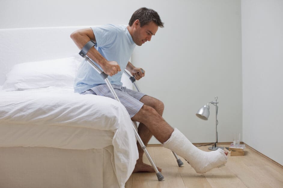 Man with a Leg Cast Sits on a Bed, Using Forearm Crutches to Steady Himself.