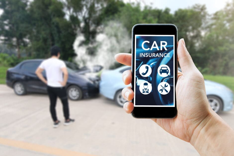 Hand Holding a Smartphone Showing a Car Insurance App; Blurred Scene of Two Damaged Cars in the Background.
