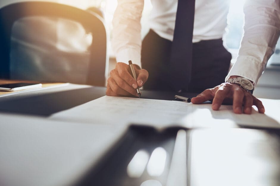 CloseUpBusinessManSigningContractMakingADealClassic - Attorneys.Media - Legal Expert Interviews & Trusted Advice Businessperson Signing a Document at a Desk, with Sunlight in the Background.