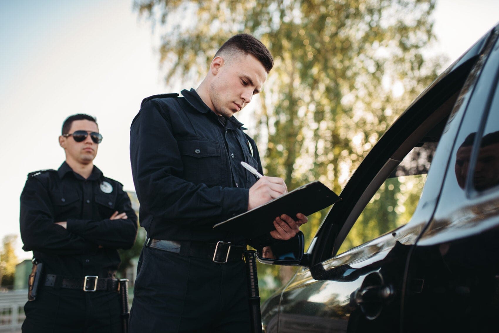 pdzff6102c5b24ea3e9201d7bd01e4a85f1 - Attorneys.Media - Legal Expert Interviews & Trusted Advice Two Police Officers in Black Uniforms Outside a Vehicle; One Writes on a Clipboard While the Other Stands Nearby in Sunglasses.